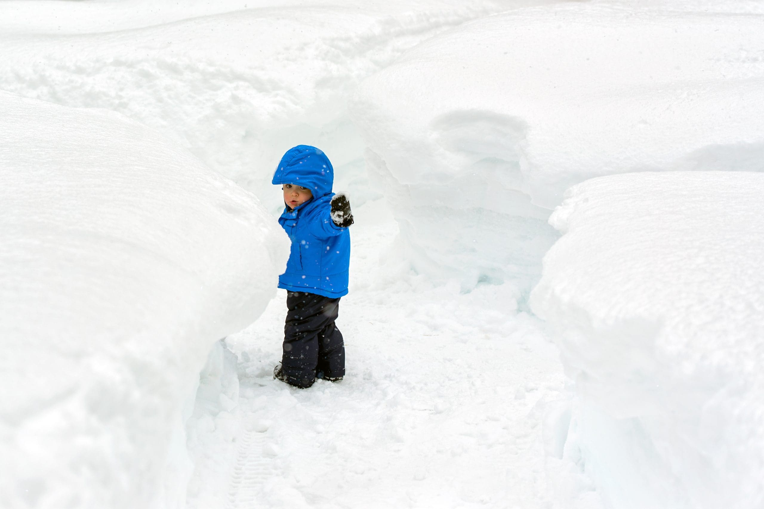 A small toddler boy points the way as he attempts navigate pathways carved in snow accumulation taller than he is. Typical winter weather within the lake effect or snow belt  regions of United States and Canada.